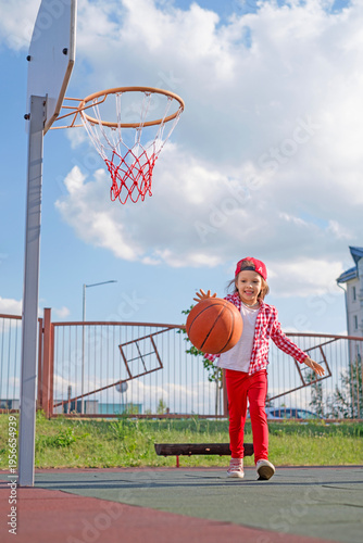 A happy little laughing girl in a baseball cap leads a basketball on the playground of a kindergarten