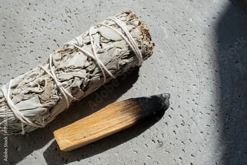 Smoldering palo santo stick and white sage bundle on ceramic plate over marble background. Spiritual energy cleansing concept. 