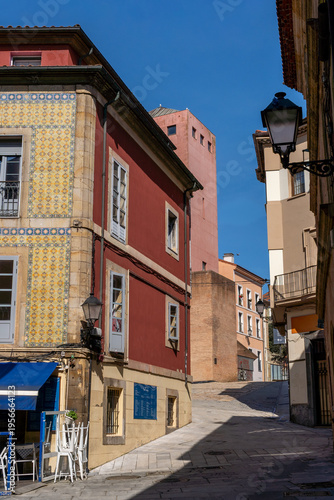 A scenic view of a narrow cobblestone street in the historic Cimadevilla district of Gijón, Spain, featuring traditional architecture with colorful tiles, old lanterns, and vibrant facades 