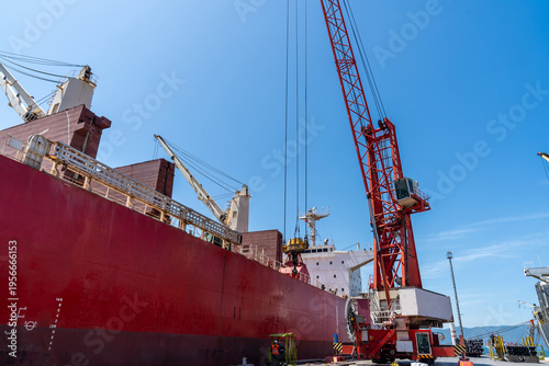 Large red cargo ship being loaded at an industrial port with a towering crane under clear blue sky. Maritime logistics, global trade, shipping industry and heavy machinery concept with copy space.