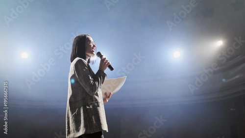 Confident businesswoman in glasses speaking into microphone while addressing audience on stage. Female presenter delivering speech at conference, seminar or corporate event under professional lighting