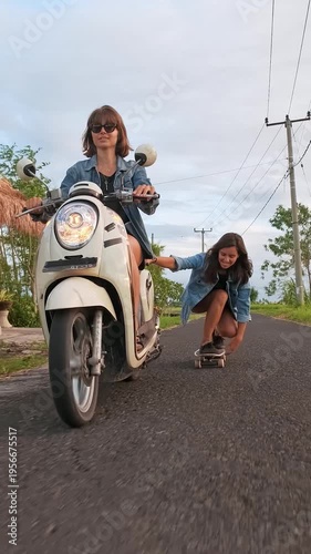 Young woman crouching on a skateboard while holding a scooter with one hand as she moves along a countryside road in Bali, Indonesia. Lush green rice paddies and a traditional building appear in the