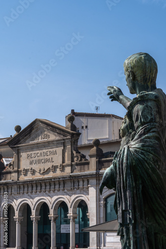 Bronze statue of Roman Emperor Octavian Augustus overlooking the historic 1928 Municipal Fish Market building in Gijon, Asturias, Spain, under a clear blue sky.