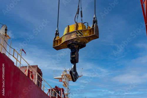 Heavy duty crane hook suspended above a cargo ship at industrial port under dramatic blue sky. Maritime logistics, shipping operations, heavy lifting and global trade concept with strong perspective.