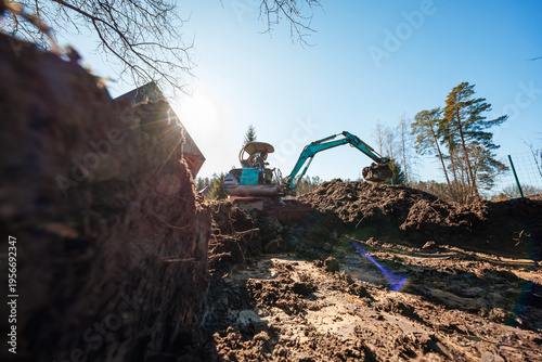 A compact teal excavator lifts wet soil from a deep trench as sunlight flares through bare branches by a sloped roof, muddy ground, wire fence, and tall pines.