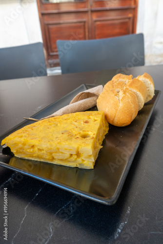 A delicious slice of traditional Spanish potato omelette (tortilla de patatas) served on a black plate with sliced bread and a small knife at an outdoor cafe. (158 characters)