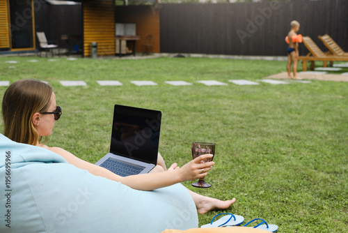 Freelancer woman works on laptop in backyard garden with wine glass while son plays in background. Concept of remote work and relaxing summer lifestyle.
