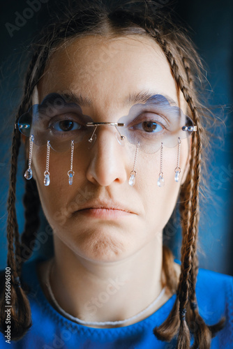 Close-up of sad young woman with braids wearing unique conceptual glasses with hanging crystal tears. Moody, artistic image for concepts of emotion, grief, and mental health.