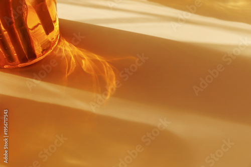 Abstract golden light refraction and elongated shadow from amber glass in warm sunlight creating minimal background with soft gradient glow and flowing reflection pattern suitable for luxury wellness