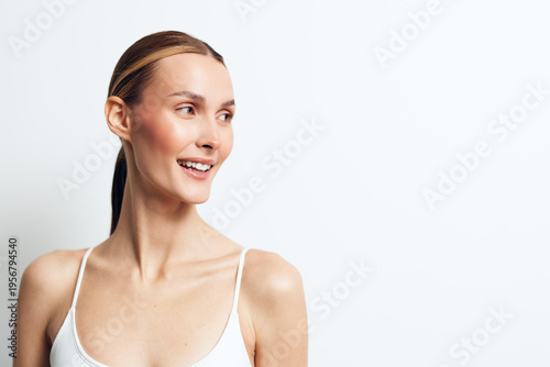 Smiling woman with clear skin and tied hair wearing a white tank top looking to the side on a white background. Concept of natural beauty, health, and happiness with soft lighting.