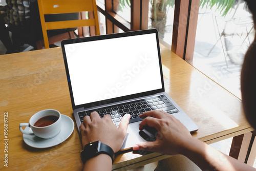 Schilderij op canvas Over the shoulder view of a person typing on a laptop with a blank white screen at a wooden table in a cafe next to a cup of coffee