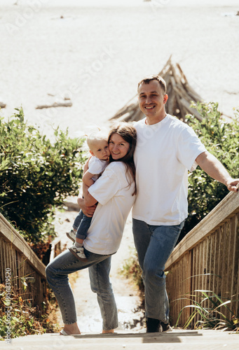 The photograph depicts a family standing on a staircase leading to the seashore. The man and woman are hugging each other, and a small child is nearby. Everyone looks happy and relaxed, enjoying their