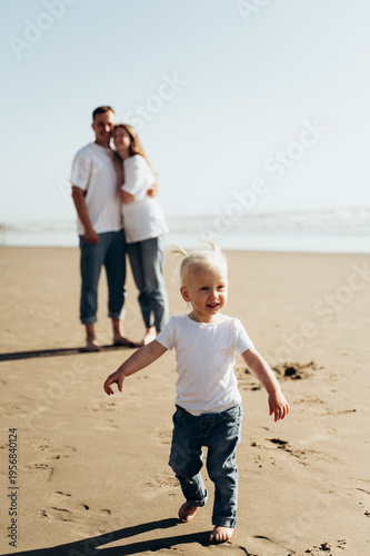 A small child walks barefoot along a sandy beach. He smiles and looks very happy. In the background, his parents stand, hugging each other and watching his child. The photograph conveys a sense of fam