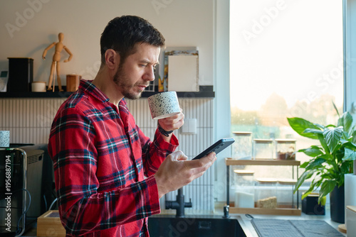 Man in red pajamas starting day with coffee and smartphone. Person drinking coffee and using mobile phone to check news at home