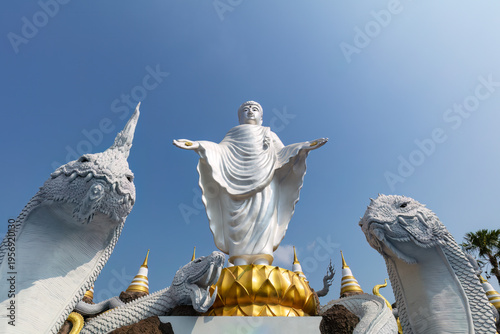 White statue of Buddha, flanked by a pair of naga serpents, at the Buddhist temple of Wat Don Yai, northeast of Bangkok, Thailand. Blue sky in the background.