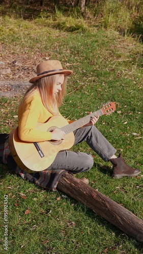 Beautiful blonde girl playing acoustic guitar and singing relaxing enjoying outdoors on sunny autumn day. Hippie woman in boho style learning and practicing playing musical instrument. Fall leaves