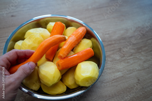 Fotografie Hand holding fresh carrot over bowl of peeled potatoes on wooden table, healthy food preparation and home cooking concept with copy space