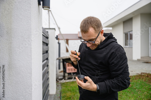 Adult man in black hoodie repairs or assembles outdoor lamp using screwdriver. Home maintenance task in backyard, DIY installation and improvement concept.