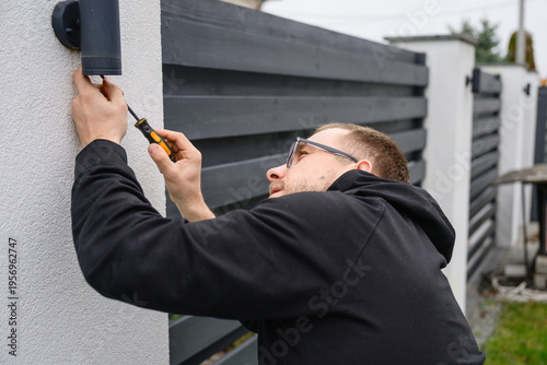 Adult man in casual clothes mounts outdoor wall lamp using screwdriver. Home improvement work, maintenance and installation in backyard of modern house.