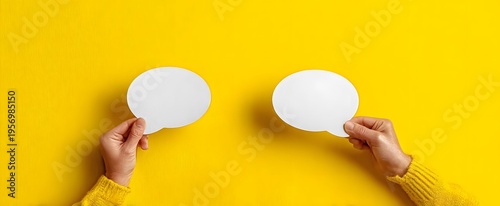 Two hands holding up blank white speech bubbles against a vibrant yellow background with soft lighting suggesting communication and interaction