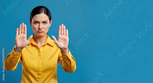 A woman with dark hair is holding her hands up in a stop gesture. She is wearing a yellow button-down shirt and has a serious expression.