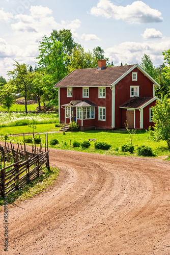 Red Scandinavian wooden house by a dirt road in the countryside in a lush green summer landscape