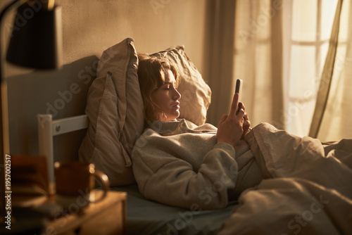Tableau sur toile Caucasian young adult woman lying in bed using smartphone, displaying neutral fa