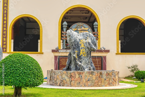 Marble rock fountain with golden Chinese characters in front of a Chinese temple facade, surrounded by trimmed bushes and stone base in a manicured courtyard.