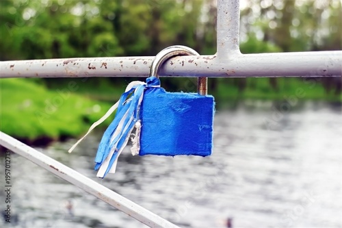 Blue padlock with fabric ribbons hangs on a rusted metal railing, set against a blurred green landscape and flowing river.