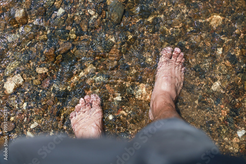 Bare foot standing in shallow clear river water over small brown rock feeling peaceful and calm enjoying relaxed outdoor nature summer stream hiking adventure
