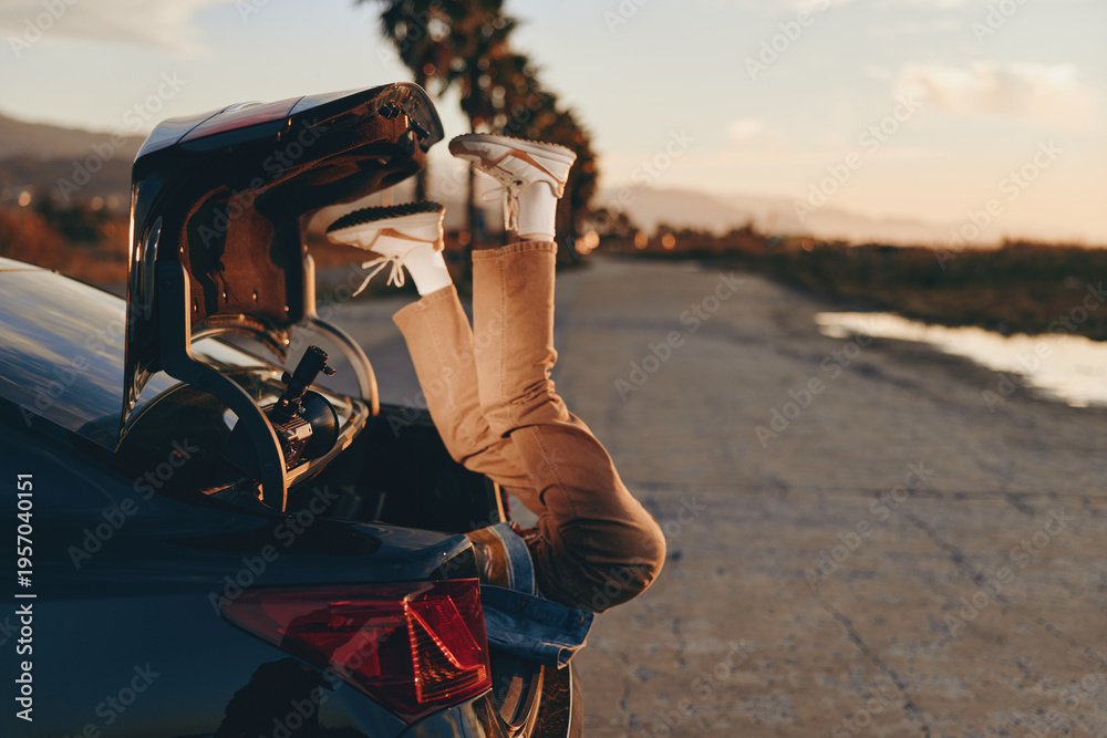 Fototapeta premium Woman wearing casual pants and white sneakers lying upside down in car trunk at sunset, relaxed lifestyle, outdoor scene with road and palm trees in background.