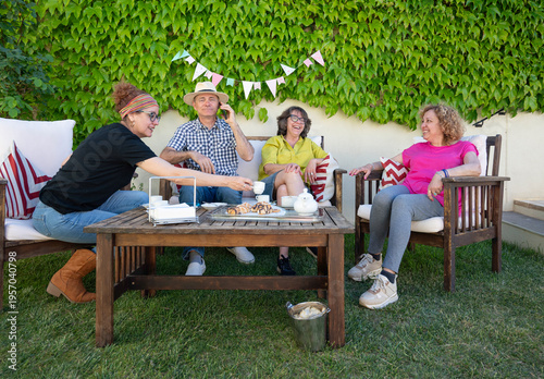 Group of middle aged friends enjoying coffee and pastries while chatting and relaxing together on sunny garden patio at home