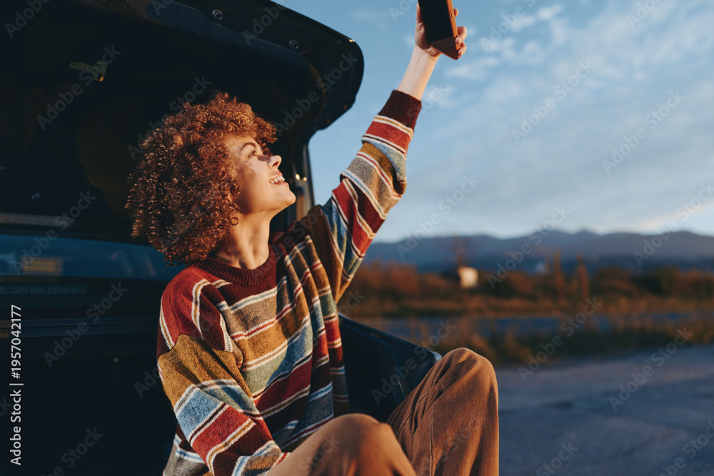 Fototapeta premium Woman smiling outdoors sitting near car trunk wearing rainbow sweater holding smartphone up enjoying lifestyle moment with natural light and clear sky background in casual relaxed pose