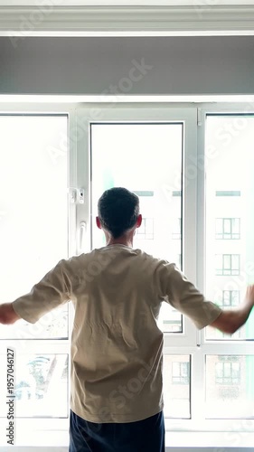 Rear view of a man waking up and opening the curtains in his modern apartment bedroom. He is looking out the window to the bright morning light