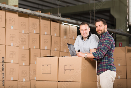 Diverse logistics team of manager and worker using laptop for inventory management, successful partners analyzing shipping data and orders among cardboard boxes in warehouse.