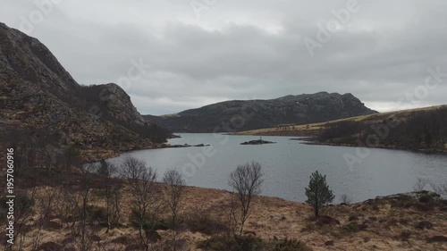 Aerial drone footage showing a mountain lake under cloudy weather conditions. The scene features calm water, surrounding mountainous terrain, and overcast skies, presenting a natural landscape with di