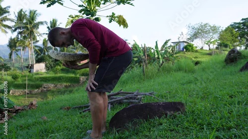 A man sits on the stone beside a papaya tree and eats rambutan 