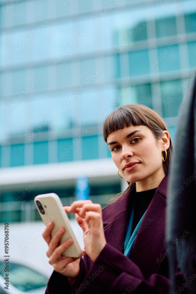 Fototapeta premium Woman standing outdoors, holding and typing on a smartphone, looking at the camera with a calm expression