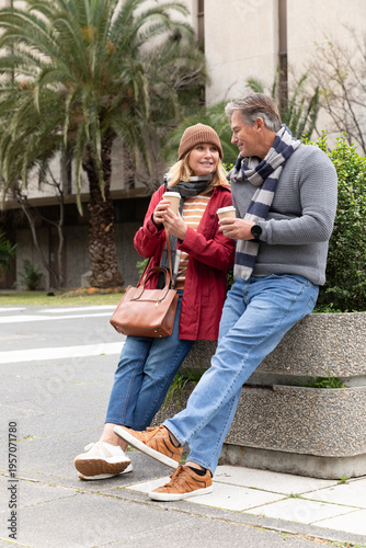 Senior couple leaning on planter bench at street, holding coffee cups, wearing beanie and scarf