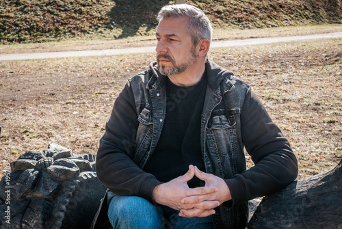 A middle-aged man sits on a carved wooden bench next to a rustic sculpture, looking to the side with a pensive expression in a sunlit park setting. Natural tones and soft shadows enhance the mood.