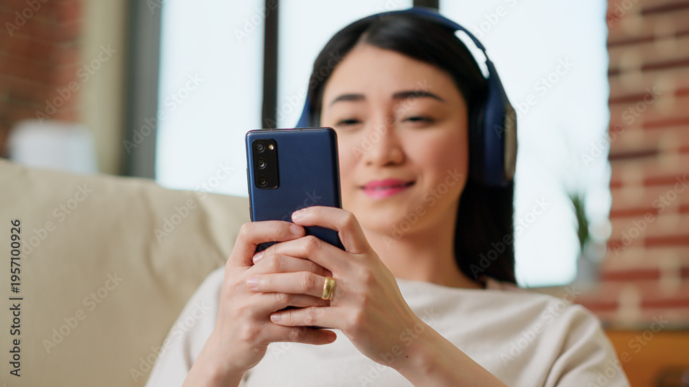 Fototapeta premium Happy young adult person chatting with friend on smartphone while working remotely. Smiling woman sending text messages on mobile phone while listening to music and doing work from home. Handheld shot