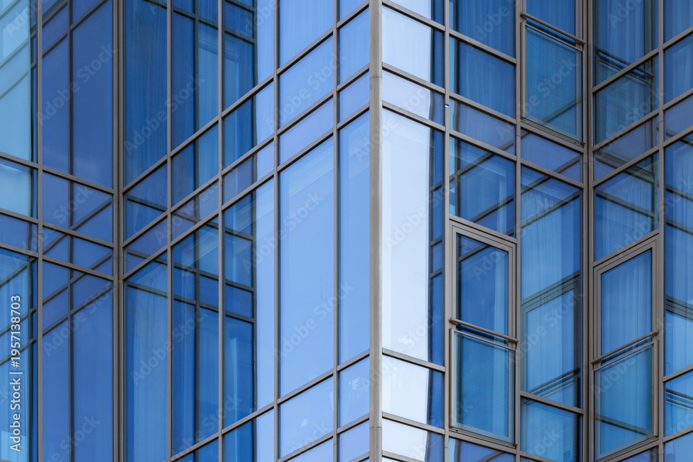 Obraz premium Modern blue glass facade of an office building with reflective windows, intersecting metal frames and geometric patterns conveying urban architecture, minimalism, transparency and contemporary design.