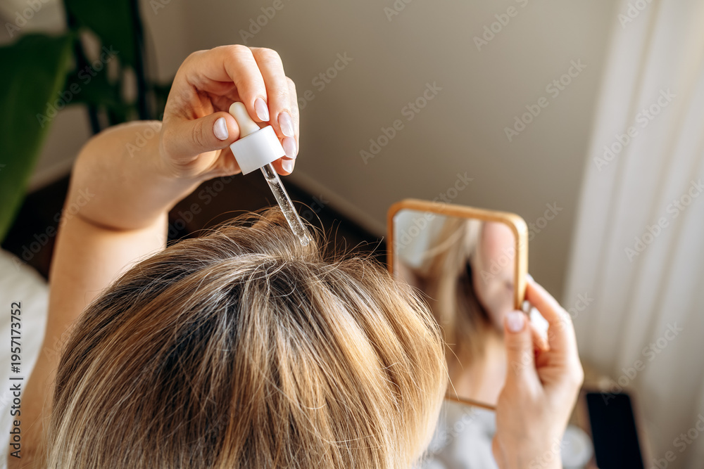Fototapeta premium Woman applying hair serum with dropper to scalp while lying on a bed, focusing on hair care and beauty routine