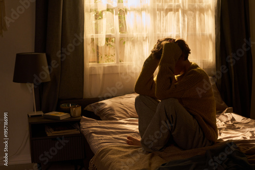 Foto Young woman sitting on bed holding head with both hands, knees drawn to chest, a