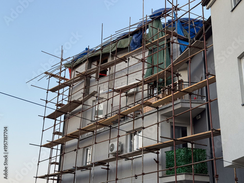 Low angle view of scaffolding around apartment facade and roof extension under tarpaulins. Residential upgrade, contractor access system, structural modification, dense neighborhood redevelopment