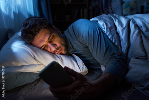 Fotografie Young man lying in bed using smartphone at night, illuminated by screen light, h