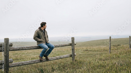 A man in a jacket and jeans sits contemplatively on a rustic wooden fence in a vast, misty green field under an overcast sky.