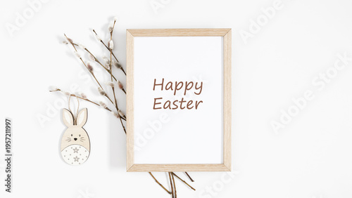 Happy Easter greeting on white background with mock up wooden frame, cute wooden bunny toy and pussy willow branches. Minimalist spring festive composition, top view, flat lay with copy space.