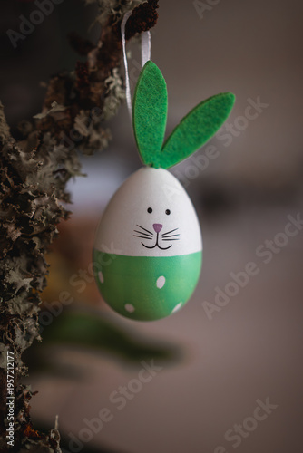 Easter bunny themed egg ornament hanging from a textured tree branch, featuring green felt ears, a hand-drawn face, and polka dots, symbolizing spring holidays and new beginnings