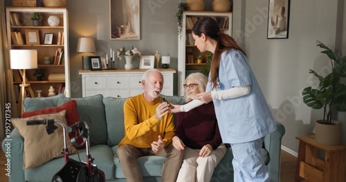 Home health nurse assists elderly couple in their living room. The nurse hands medication to the man while the woman looks on. The scene takes place in a warm home environment.